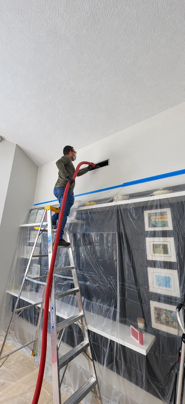 Technician cleaning a wall duct opening above furniture protected with plastic