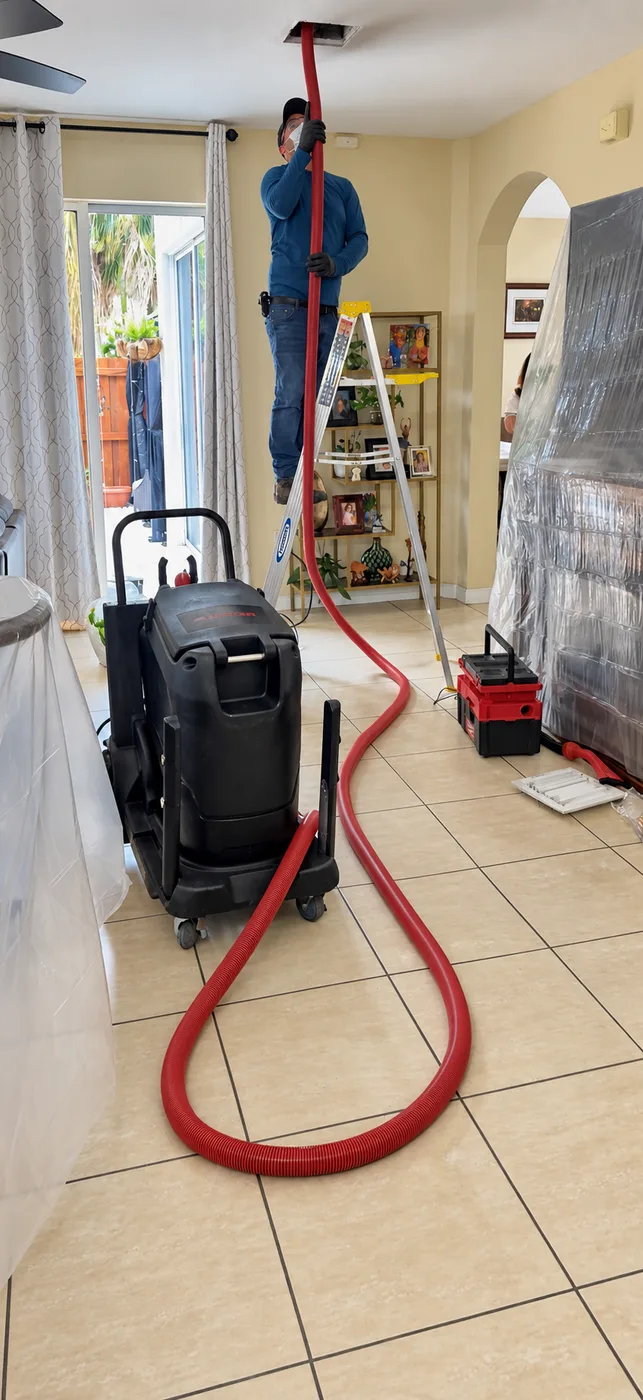 Technician cleaning a ceiling duct with a red hose and portable cleaning machine